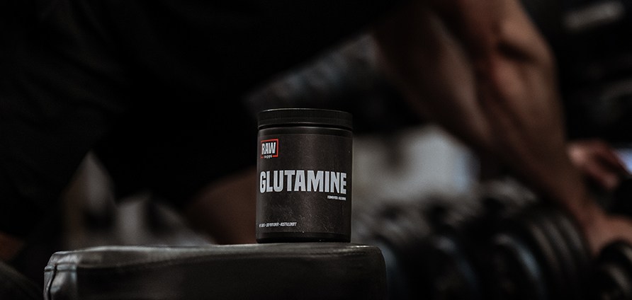 RAW SUPPS Glutamine i fokus p en bnk, med en person i bakgrunden som tar en hantel frn ett hantelrack.